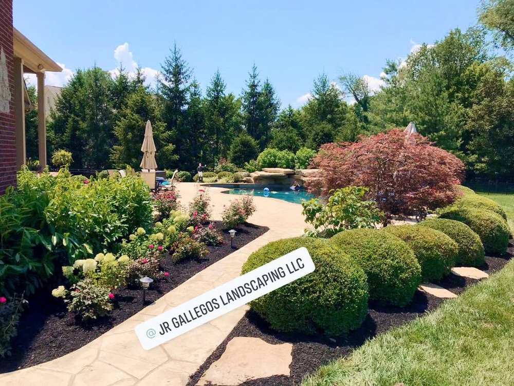 A stone path curving past deep planted beds with clipped shrubs and a red Japanese maple, running down to a swimming pool behind a large house.